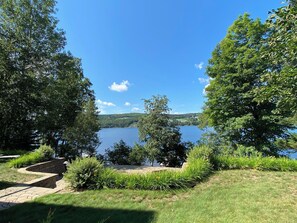 A lakeside view with a stone pathway leading to a seating area, surrounded by lush greenery and trees.