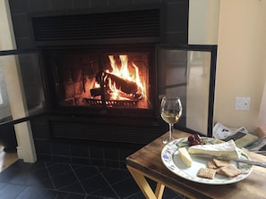 Une cheminée avec une porte en verre, une table sur laquelle se trouve une assiette de fromage et de crackers, et un verre de vin.
