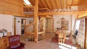 Un intérieur spacieux en bois, avec un escalier en colimaçon central, un coin repas et un salon agrémenté d’un fauteuil en cuir rouge.