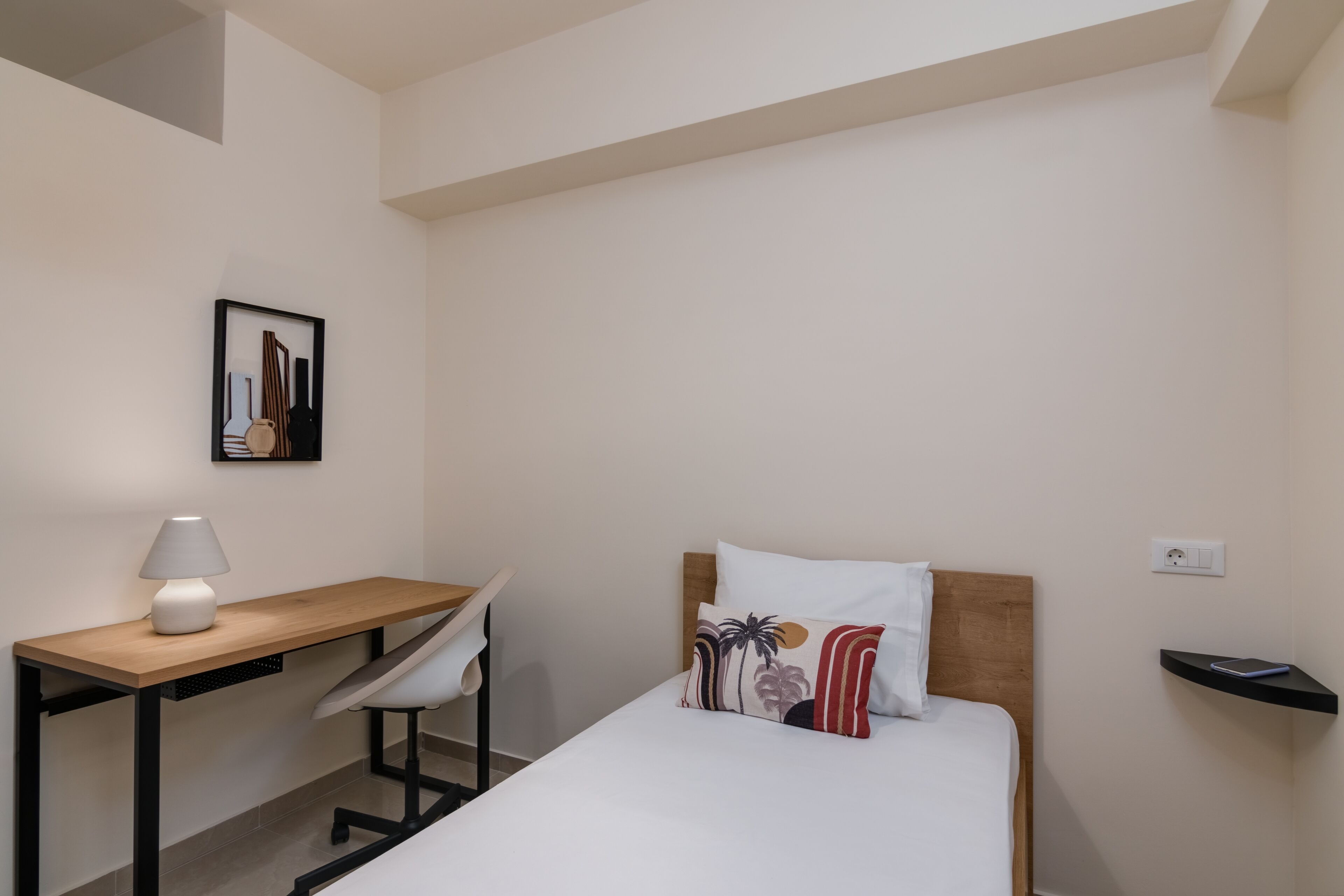Una habitación pequeña y ordenada con una cama, un escritorio y una silla. Encima del escritorio, hay un cuadro enmarcado en la pared.