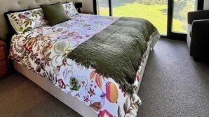 Une chambre dotée d’un grand lit, offrant une vue sur un paysage verdoyant grâce à de grandes fenêtres du sol au plafond, et comprenant un petit coin salon.