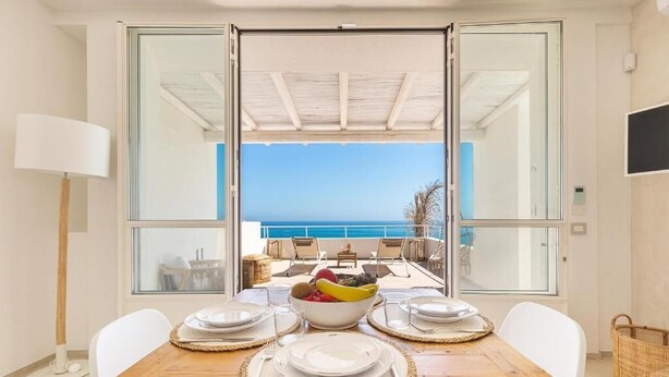 A dining area with a table set for four, a bowl of fruit, and a view of the sea through large glass doors. - Palma Residence Salento (Melendugno)