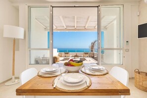 Comedor con una mesa dispuesta para cuatro, un frutero y vistas al mar a través de puertas de vidrio amplias.