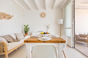 Un comedor moderno con una mesa de madera dispuesta para cuatro, sillas blancas, un sofá blanco, una planta en maceta y un adorno en la pared.