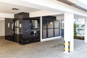 Ingresso di un edificio moderno con una porta in marmo nero, un ascensore in vetro e un cartello con indicazione di parcheggio.