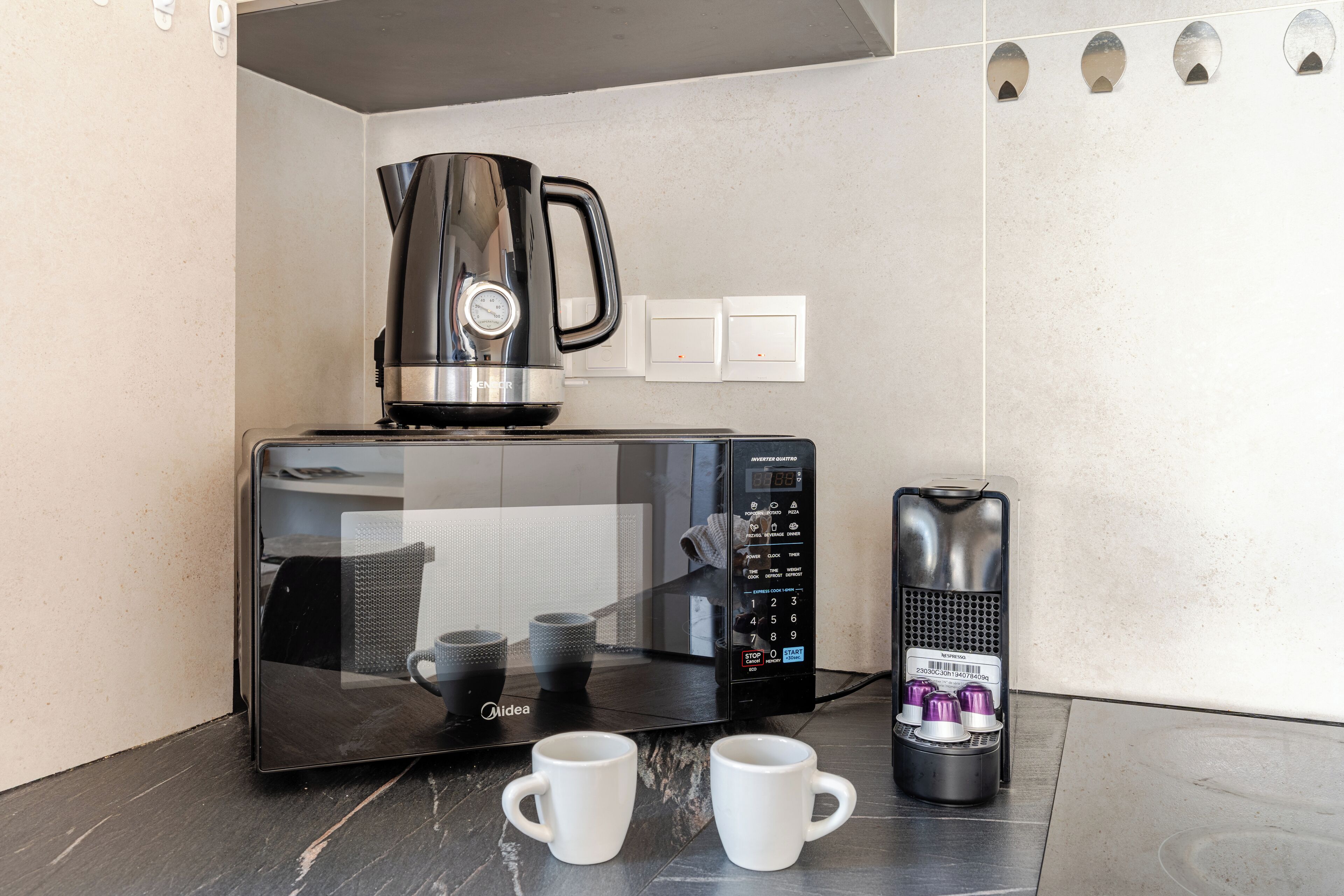 Uma cozinha compacta com micro-ondas, cafeteira e duas canecas brancas em bancada escura.
