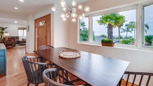 A dining area with a wooden table and chairs, a hanging light fixture, and a view of palm trees through large windows.