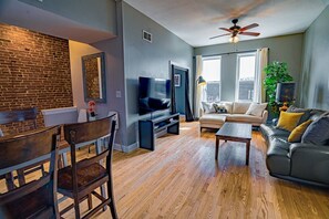 A modern living room with a brick wall, a television, a sofa, and a dining area with chairs. - Classy 3BR King Apt in The Grove (St. Louis)