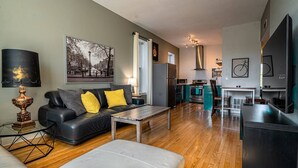 Ein modernes Wohnzimmer mit einer schwarzen Ledersofa, einem Glastisch und einem grauen Hocker. Im Hintergrund ist eine Küche mit Herd, Backofen und Kühlschrank zu sehen.