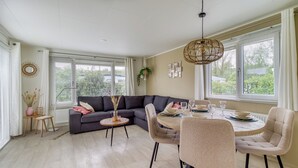 Una sala de estar moderna con un sofá seccional gris oscuro, una mesa de comedor redonda con sillas color beige y un ventanal con cortinas blancas.