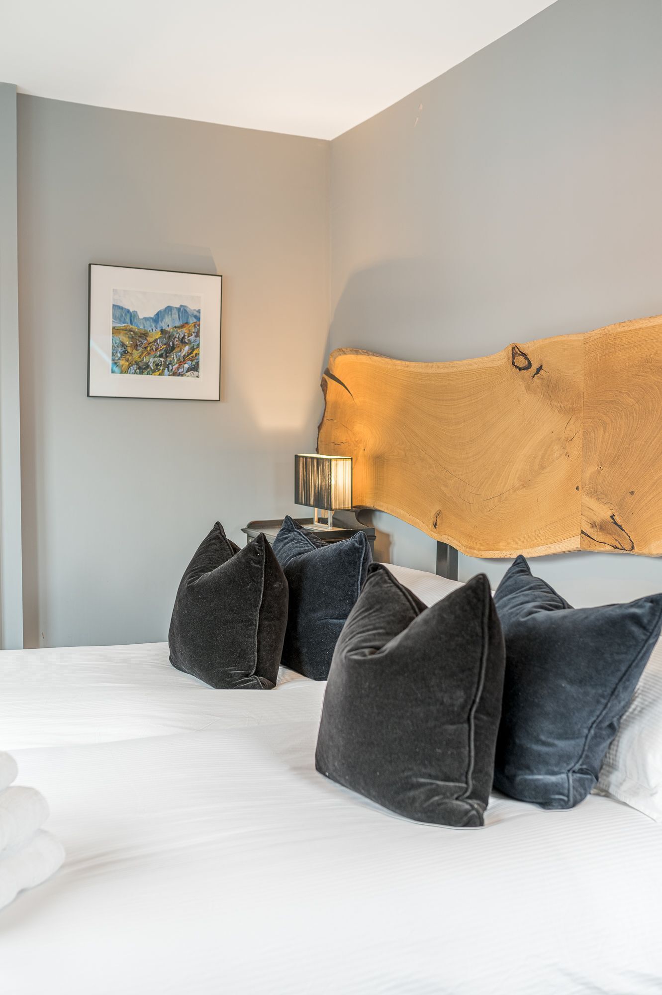 A bedroom with a bed, pillows, a nightstand with a lamp, a framed painting, and a large wooden headboard.