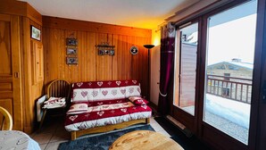 A cozy room with a wooden wall, a sofa with a heart-patterned blanket, a wooden table, and a view of a building outside. - Cozy T2 Apartment with Terrace, 5 pers, Les Saisies (Les Saisies)