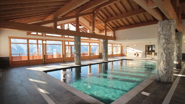 En indendørs swimmingpool med stensøjler, træbjælker og store vinduer med udsigt til bjergene.