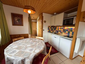 A compact kitchen with a microwave, sink, and white cabinets. There's a dining table with a patterned tablecloth and chairs. A wooden ceiling and a green curtain are also visible.