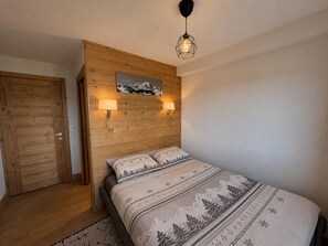 A bedroom with a wooden headboard, a bed with patterned bedding, two wall-mounted lamps, and a ceiling light fixture. - New apartment 50m², 2 bedrooms, WIFI, covered parking (Les Saisies)
