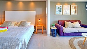 A modern bedroom with a large bed, bedside table with a lamp, a purple sofa, and three framed pictures on the wall. - Spacious Studio with View and Pool in Mont Vernon (Saint-Martin)