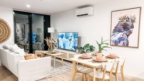 A modern living room with a white sofa, a dining table set for four, and a television on the wall. - Luxury Apartment in Canberra CBD (Reid)