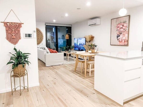 Una sala de estar moderna con un sofá blanco, un comedor con mesa y sillas de madera, una isla de cocina y un ventanal con vista a la ciudad.