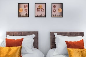 Ein Hotelzimmer mit zwei Betten, jedes mit weißer Bettwäsche und orange-gelben Kissen. Darüber an der Wand befinden sich drei gerahmte Bilder, die jeweils eine Stadtansicht bei großem Mond zeigen.