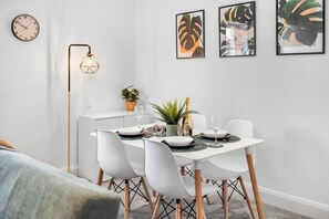 Ein Essbereich mit einem weißen Tisch, gedeckt für vier Personen, weißen Stühlen, einer Stehlampe und gerahmten Pflanzenmotiven an der Wand.