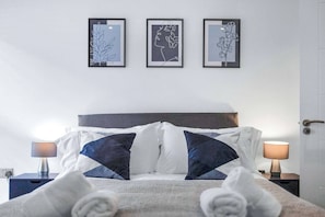 A neatly made bed with white and dark blue pillows, a headboard, and a nightstand with a lamp. Three framed artworks are hung on the wall above the bed.