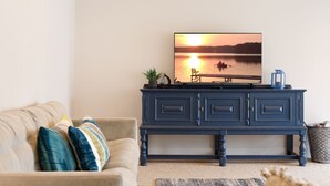 A living room with a beige sofa, a dark blue cabinet, and a television displaying a sunset over a lake. - Green Lake Central Interlochen | Firepit (Interlochen)