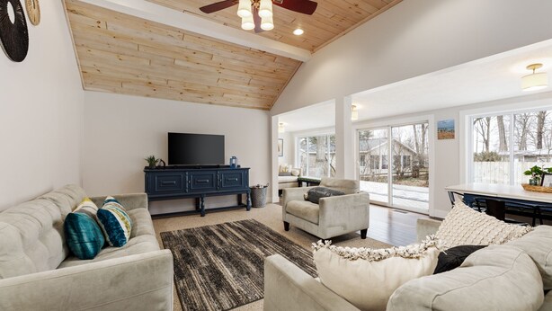 A spacious living room with a high ceiling, a ceiling fan, a flat-screen TV, a dark blue cabinet, and a large window offering a view of the outdoors. - Green Lake Central Interlochen | Firepit (Interlochen)