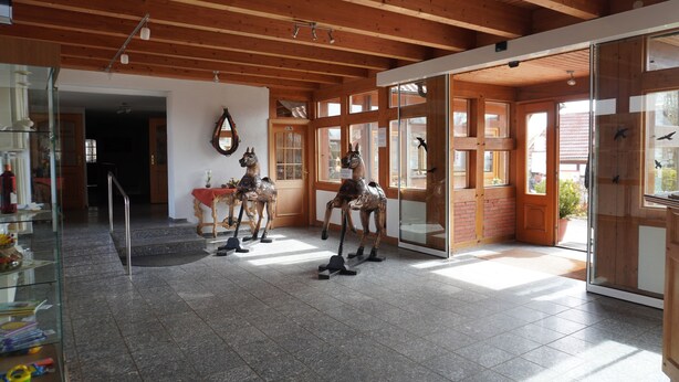A spacious room with a high ceiling, wooden beams, and large windows. There are three horse statues on the floor, and a glass door leading outside.