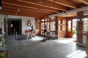 A spacious room with a high ceiling, wooden beams, and large windows. There are three horse statues on the floor, and a glass door leading outside. - Alcor Hotel Feriendorf an der Ostsee (Klütz)