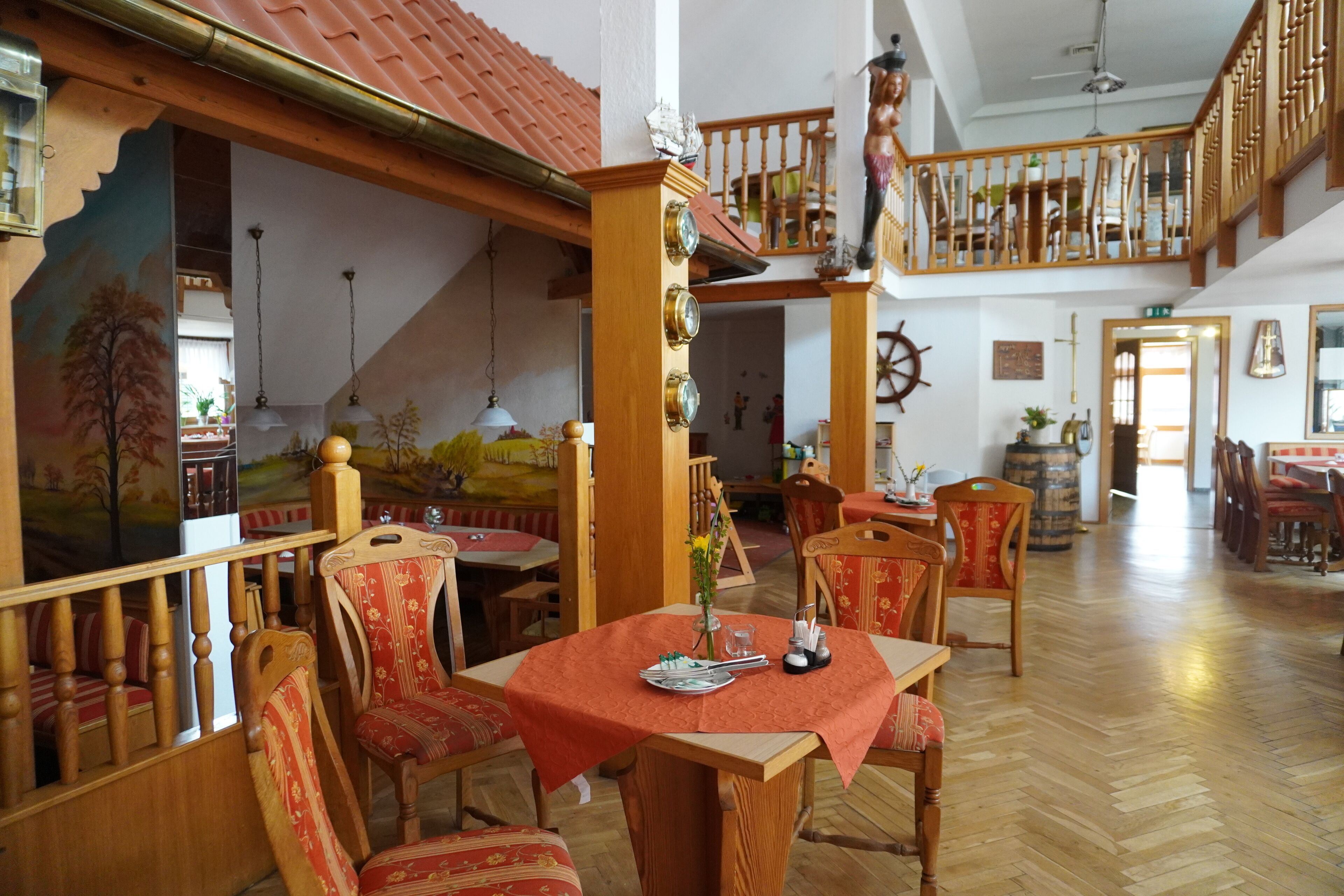 Uma sala de jantar com móveis de madeira e toalha de mesa vermelha, uma escada com corrimão de madeira e um mural na parede.