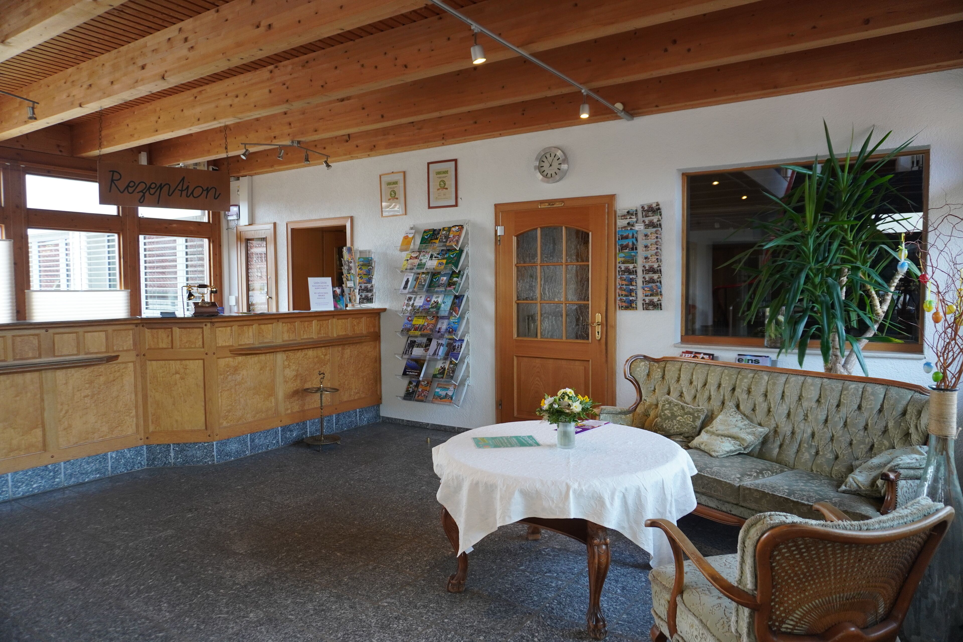Un espace d’accueil comprenant un comptoir de réception en bois, une table ronde recouverte d’une nappe blanche, un canapé confortable et une grande plante en pot.