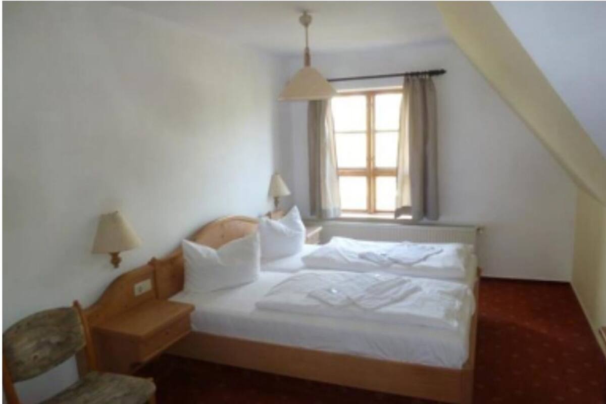 Habitació senzilla i neta amb un llit individual, una cadira, una taula de nit, una làmpada i una finestra amb cortines.