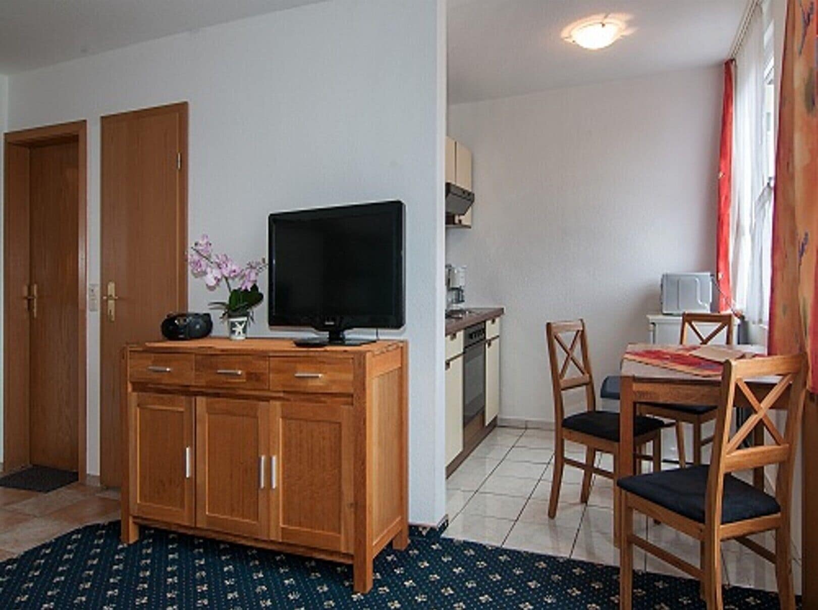 Een kamer met een houten kast, een televisie, een eettafel met stoelen, een keukenhoek en een kamerbreed tapijt met een patroon.