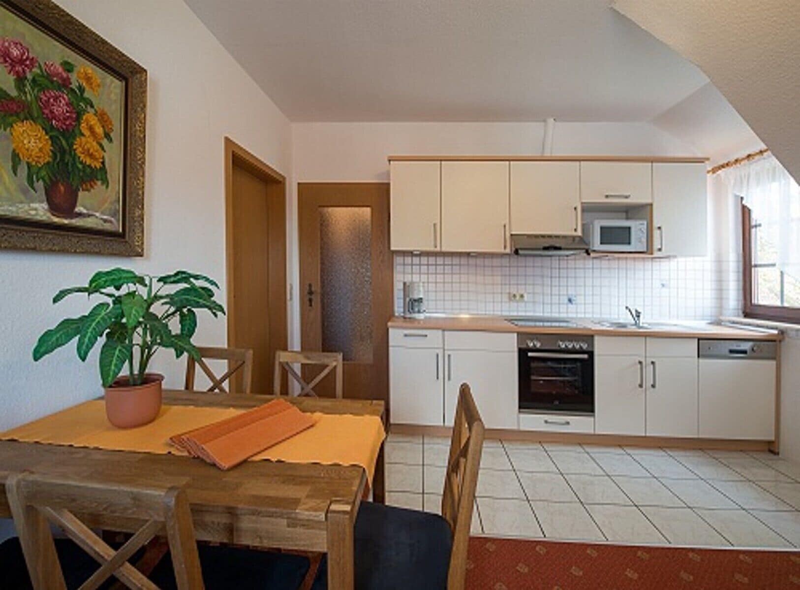 A compact kitchen with white cabinets, a microwave, and a dishwasher. There's a dining table with chairs and a potted plant.