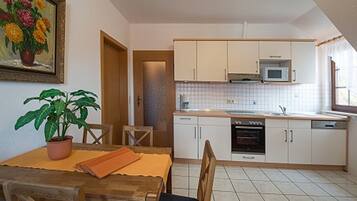 A compact kitchen with white cabinets, a microwave, and a dishwasher. There's a dining table with chairs and a potted plant.