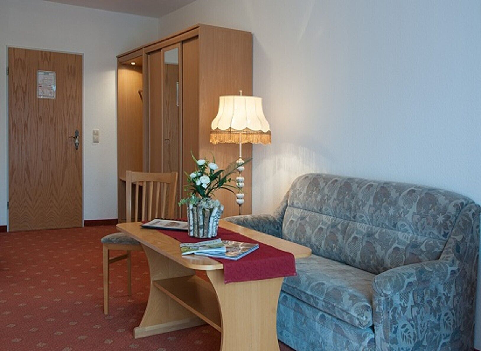 A room with a patterned sofa, a wooden table with a vase of flowers, a lamp, and a wooden door.