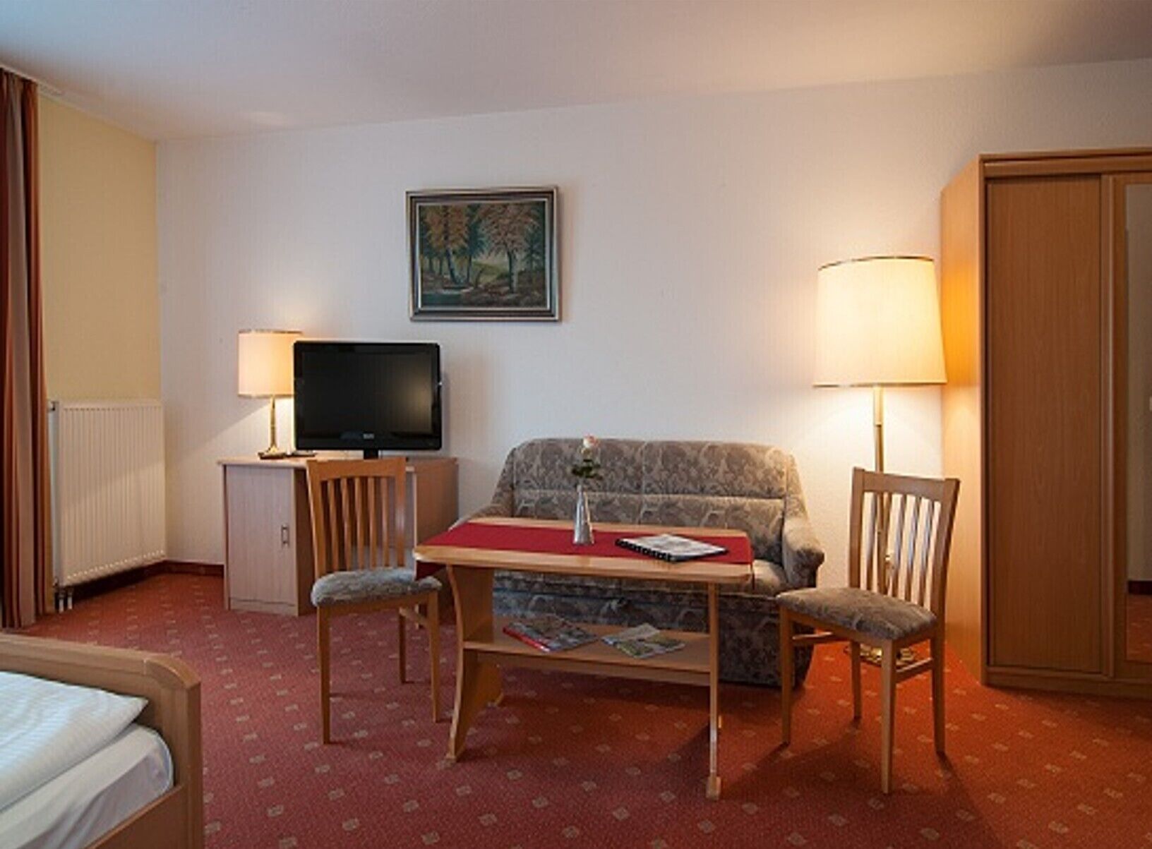 Una habitación de hotel con cama, televisión, sofá, mesa con mantel rojo, dos sillas, lámpara, un cuadro en la pared y armario.