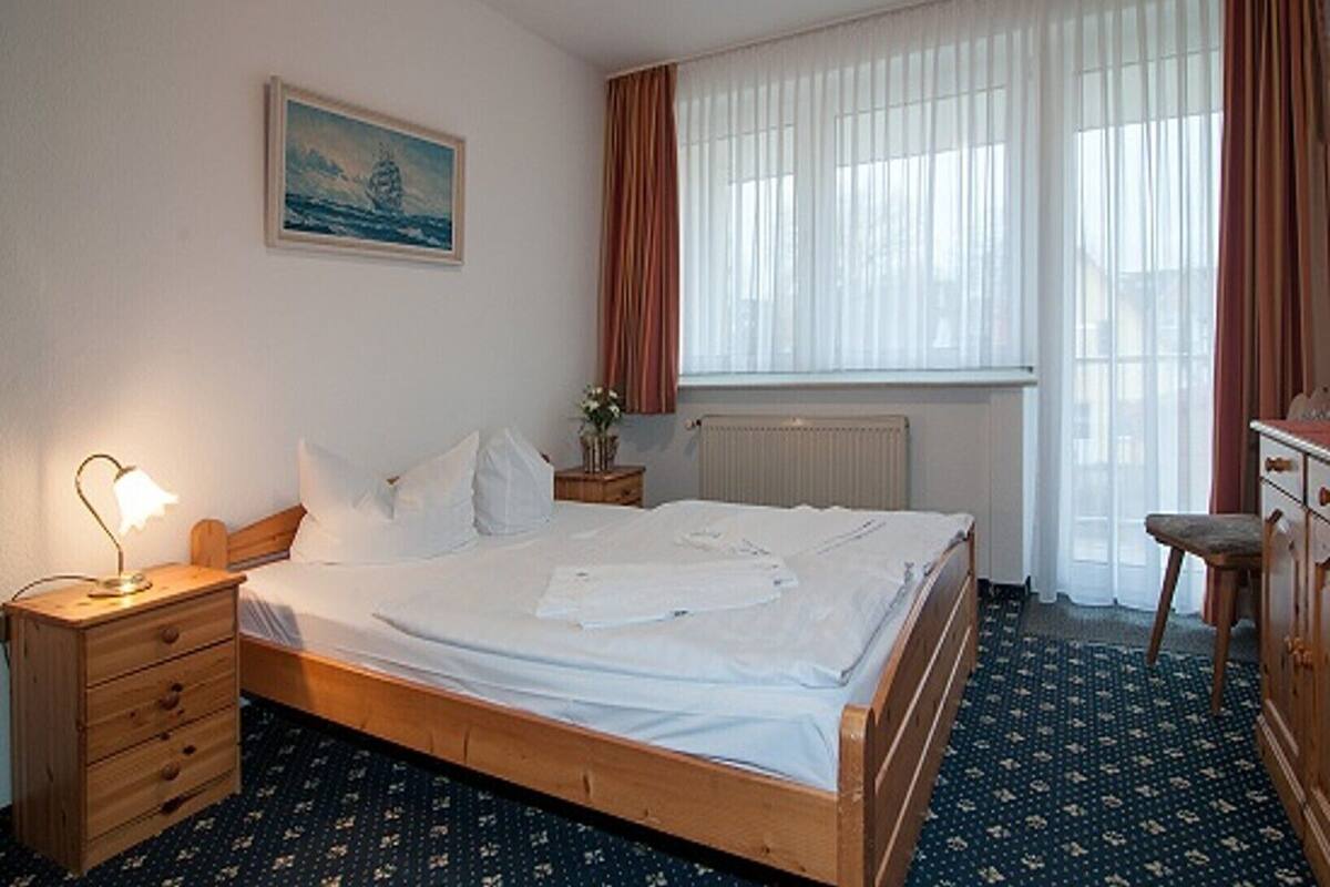 Habitació d'hotel amb llit de fusta, una nitòria amb làmpada, un prestatge i una finestra amb cortines.
