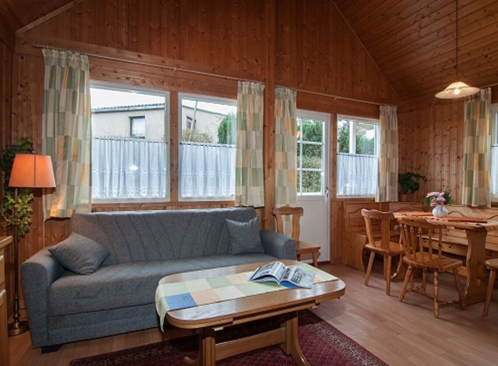 A cozy wooden cabin interior with a sofa, dining table, chairs, and large windows.