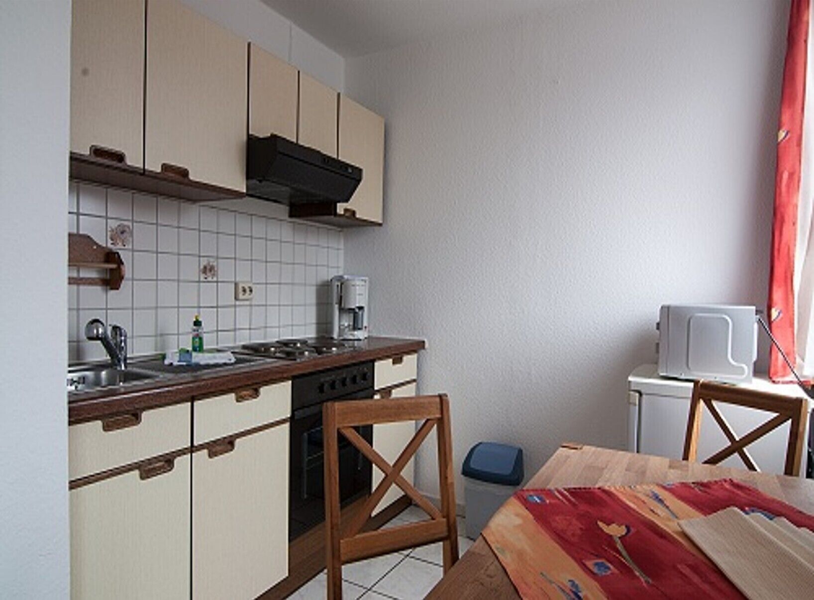 A compact kitchen with a stove, sink, and microwave. There is a small trash bin and a table with a placemat.