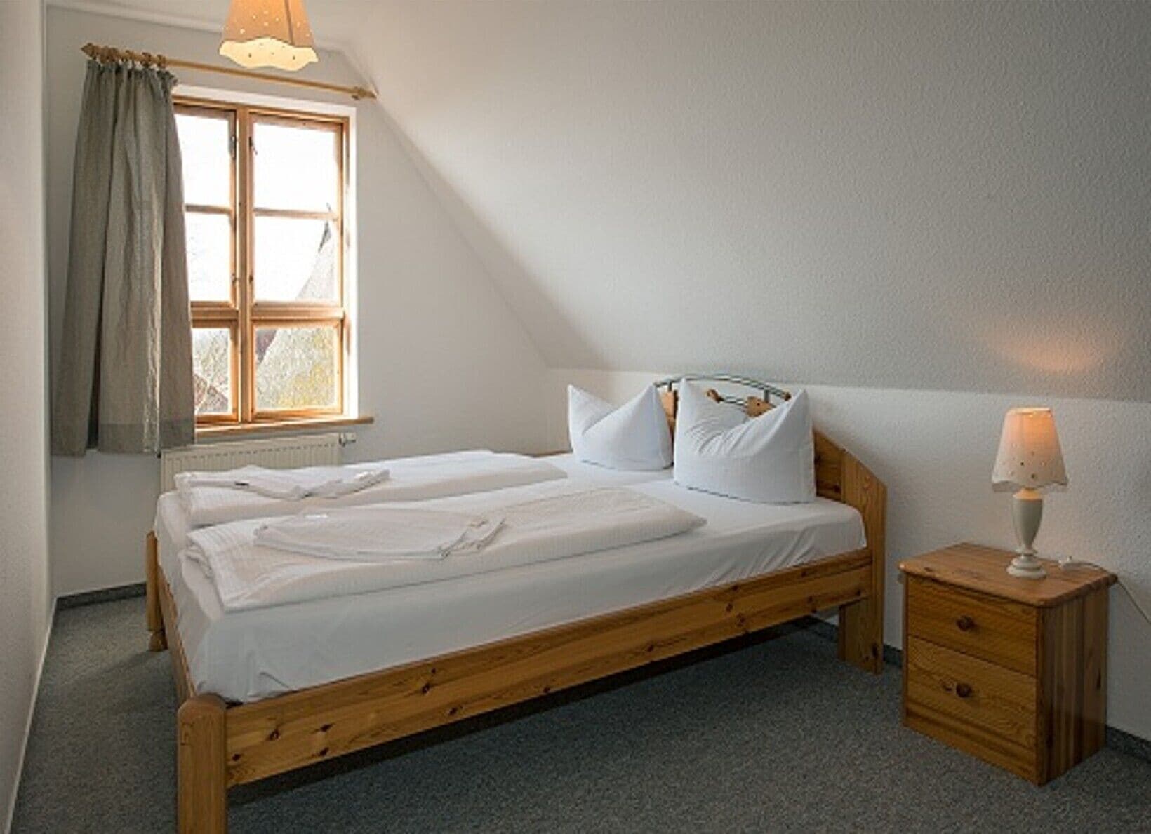 A single-bed room with a wooden frame, white bedding, a bedside table with a lamp, and a window with curtains.
