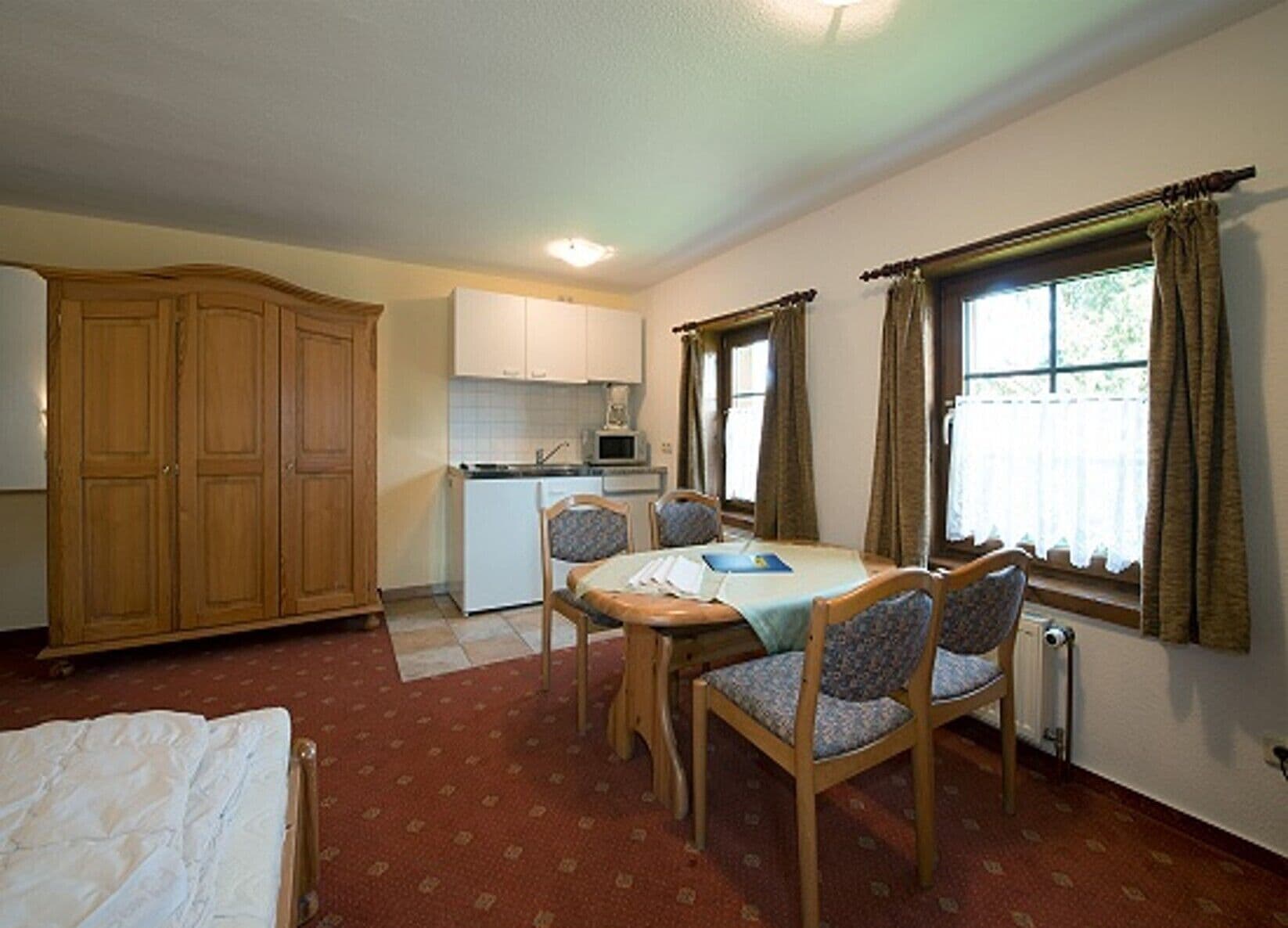 A compact room with a dining table, four chairs, a bed, a kitchenette, and a large wardrobe.