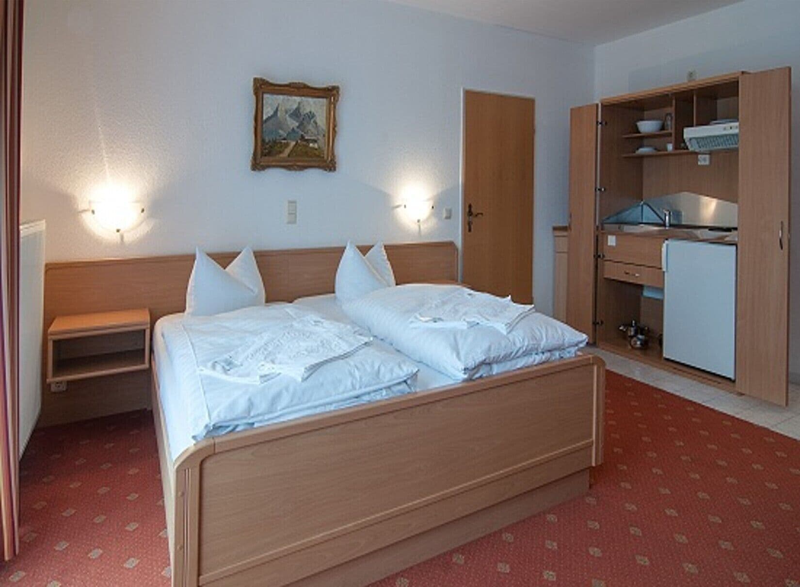 A hotel room with a wooden bed, a nightstand, a wardrobe, and a small desk with a chair.