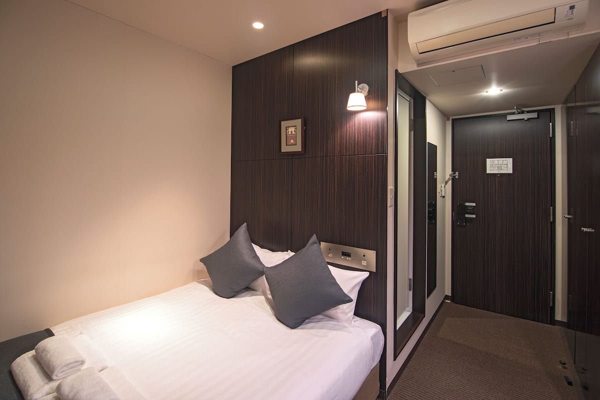 Quarto de hotel com cama, almofadas, luminária na parede, ar condicionado e uma porta.