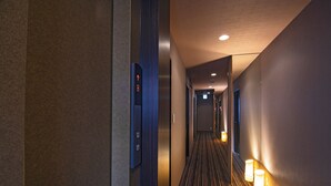Un couloir d'hôtel avec un sol recouvert de moquette, des luminaires fixés au mur et une porte dotée d'un panneau de boutons pour l'ascenseur.