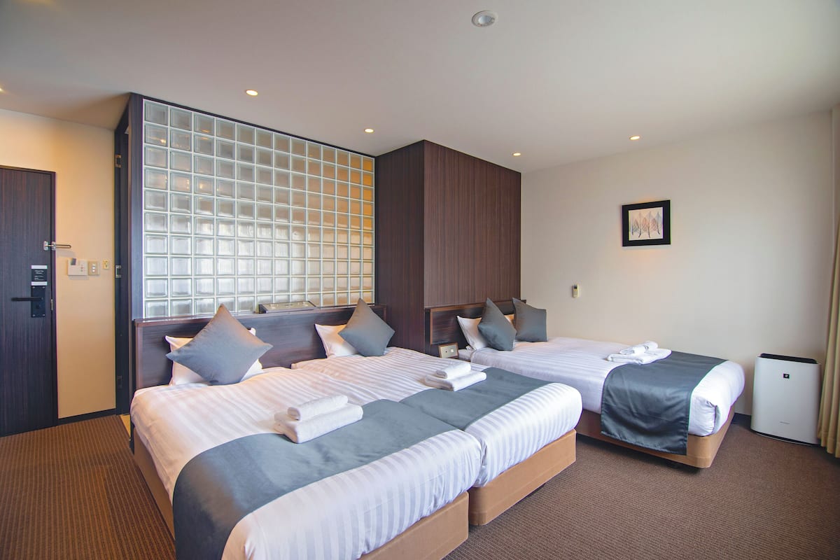 Quarto de hotel com duas camas, uma janela grande com vista, uma mesa de cabeceira e um pequeno ar condicionado.