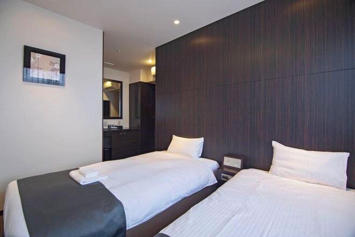 A hotel room with two beds, a dark wood panel wall, a framed picture on the wall, and a small bedside table with a lamp.