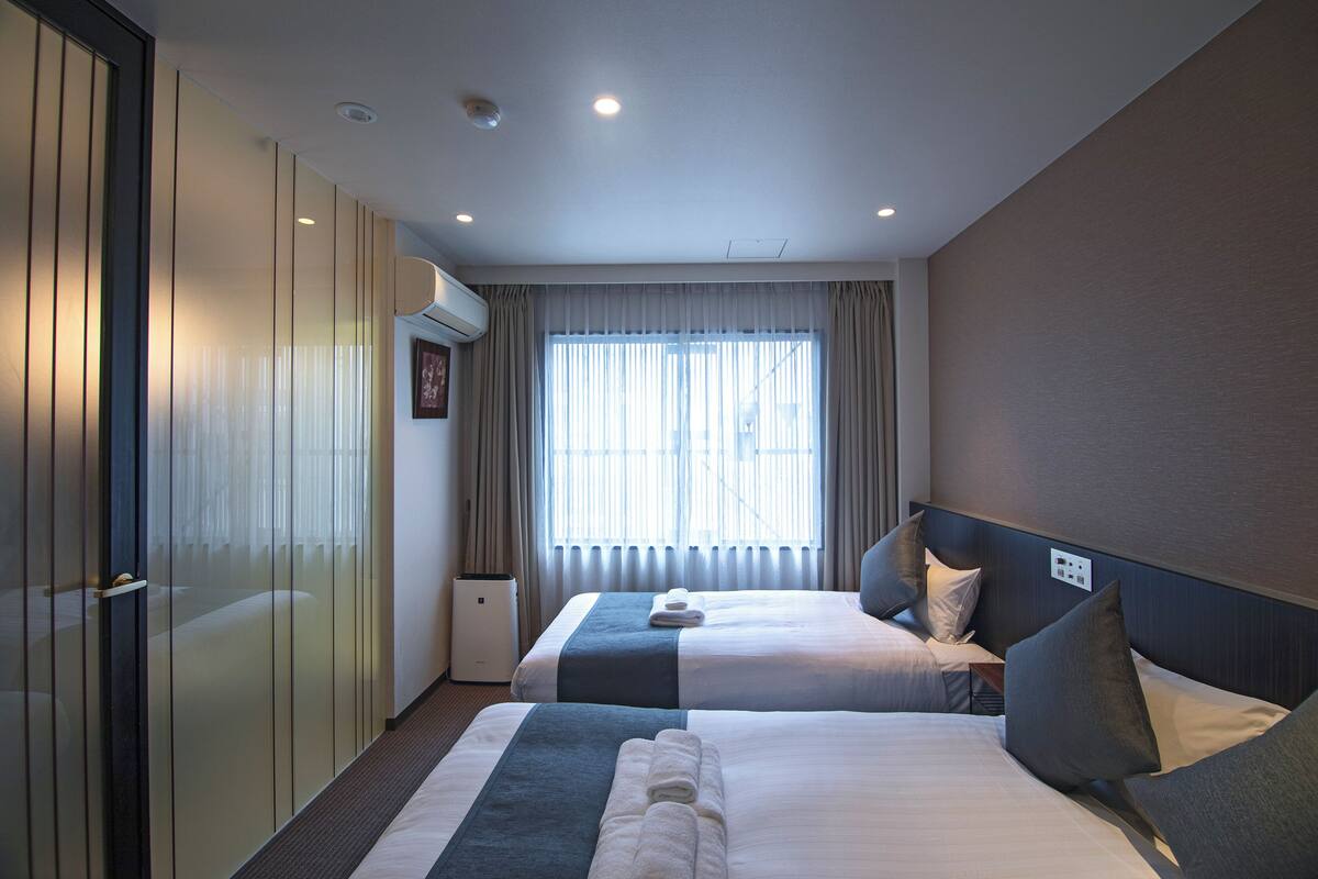 Quarto de hotel com duas camas, um guarda-roupa, janela com cortinas e uma lâmpada no teto.