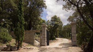 Une entrée fermée par un portail, avec des piliers en pierre et un portail en métal, le tout entouré d’arbres et bordé d’un chemin de gravier.