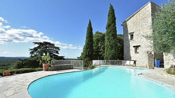 Une piscine aux bords recouverts de pierres, entourée d’un bâtiment en pierre et de grands arbres.